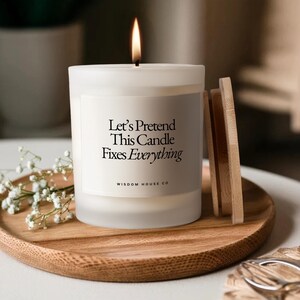 May include: A lit white candle with the text "Let's Pretend This Candle Fixes Everything" on a wooden tray. The candle has a wooden lid and is next to a sprig of white flowers. The candle is from Wisdom House Co.