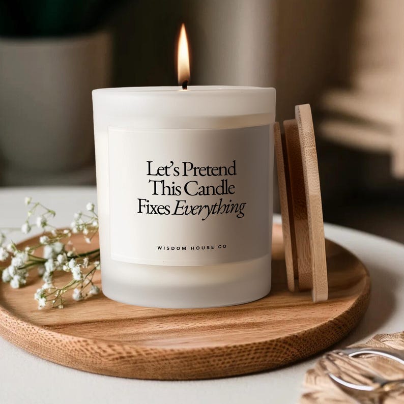 May include: A lit white candle with the text "Let's Pretend This Candle Fixes Everything" on a wooden tray. The candle has a wooden lid and is next to a sprig of white flowers. The candle is from Wisdom House Co.