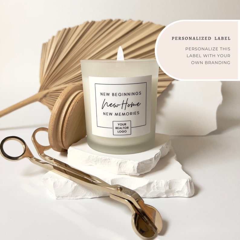 K&ouml;nnte beinhalten: Wei&szlig;e, gefrostete Glaskerze mit einem wei&szlig;en Etikett, auf dem "NEW BEGINNINGS new Home NEW MEMORIES YOUR REALTOR LOGO" steht. Die Kerze steht auf einer wei&szlig;en, felsartigen Oberfl&auml;che mit einem goldenen Kerzenl&ouml;scher.