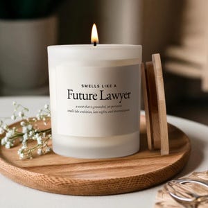 May include: A white candle with the text "SMELLS LIKE A Future Lawyer" on a wooden tray. The candle has a wooden lid and the text "a scent that is grounded, yet persistent smells like ambition, late nights, and determination."