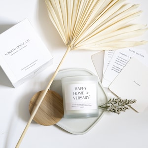 May include: White candle with a frosted glass jar and a black label that reads "Happy Home-A-Versary" wishing you another year full of memories and making a house a home.  The candle is on a white tray with a wooden lid and a small bouquet of white flowers.
