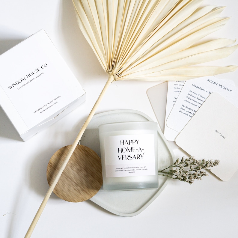 May include: White candle with a frosted glass jar and a black label that reads "Happy Home-A-Versary" wishing you another year full of memories and making a house a home.  The candle is on a white tray with a wooden lid and a small bouquet of white flowers.