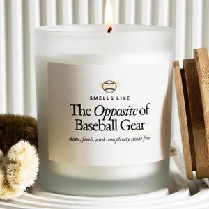 May include: A lit white candle in a frosted glass jar with a wooden lid. The label reads "Smells Like The Opposite of Baseball Gear" with the text "clean, fresh, and completely sweat-free". A baseball graphic is above the text.