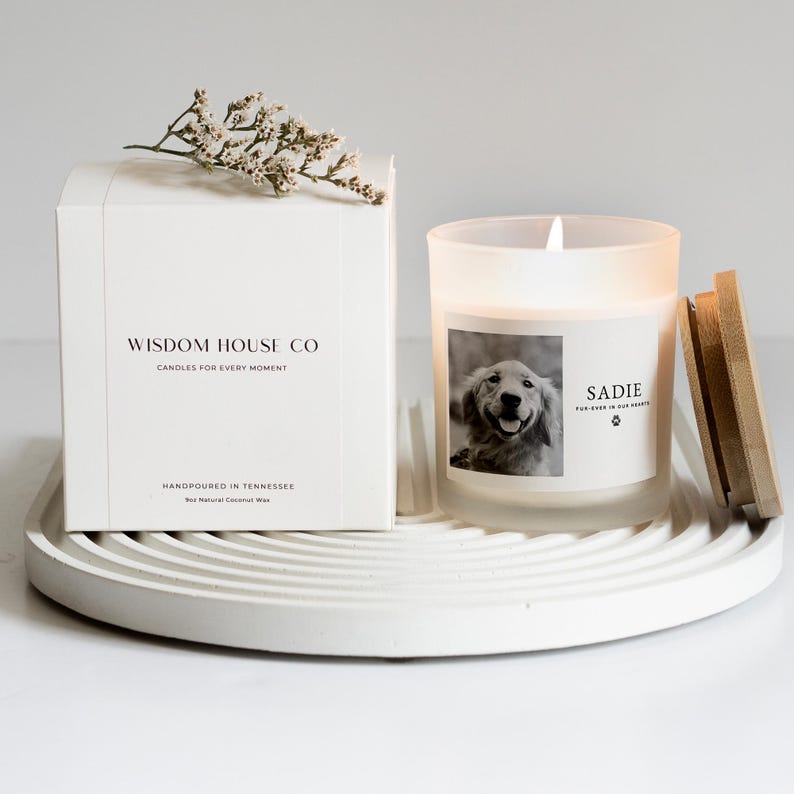 May include: A white box with the text "WISDOM HOUSE CO CANDLES FOR EVERY MOMENT" and a lit candle in a frosted glass. The candle has a photo of a dog and the name "SADIE". The candle sits on a white, circular tray.