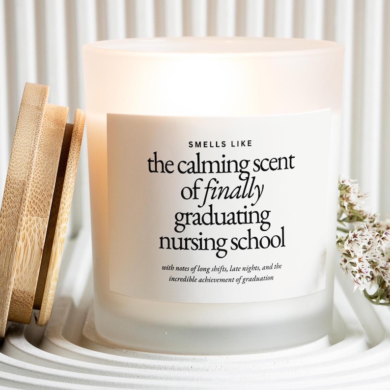 Może przedstawiać: Świeca z matowego szkła z drewnianą pokrywką, z napisem "Smells Like the calming scent of finally graduating nursing school". Etykieta zawiera r&oacute;wnież informacje o długich zmianach i p&oacute;źnych nocach. Świeca stoi na białej tacy.