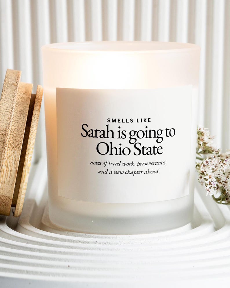 Puede incluir: Una vela de vidrio esmerilado con tapa de madera. La vela tiene una etiqueta blanca con el texto "Smells Like Sarah is going to Ohio State". El texto de abajo dice "notes of hard work, perseverance, and a new chapter ahead".