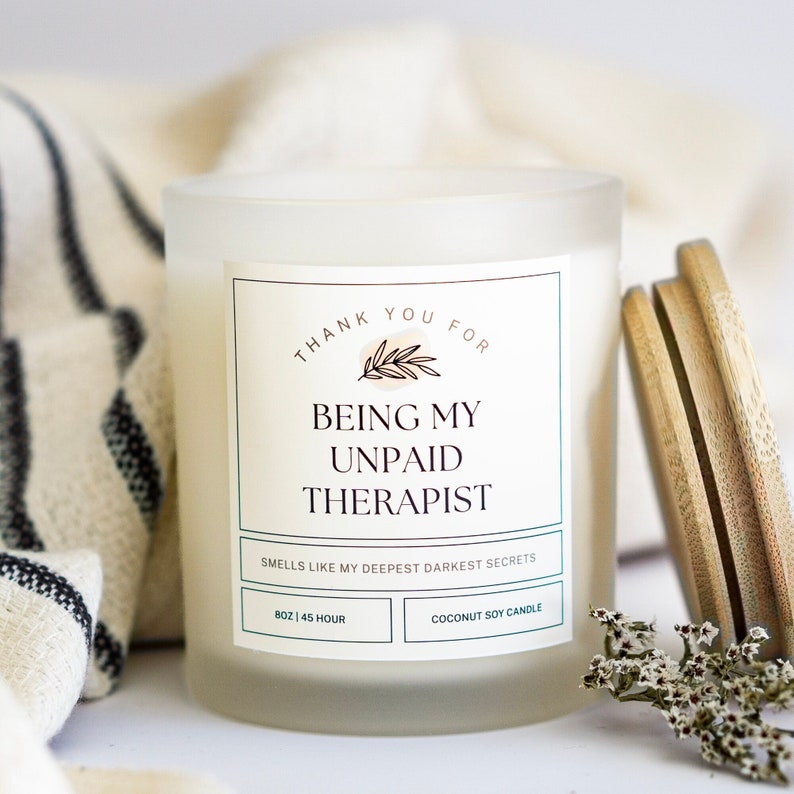 May include: White frosted glass candle with a label that reads "Thank you for being my unpaid therapist. Smells like my deepest darkest secrets. 8 oz | 45 hour. Coconut soy candle."