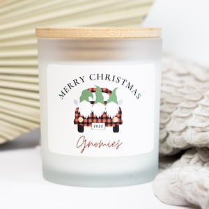 May include: White frosted glass candle jar with a wooden lid. The label features a red and black checked truck with three gnomes in the back. The text on the label reads "Merry Christmas Gnomes 2022".