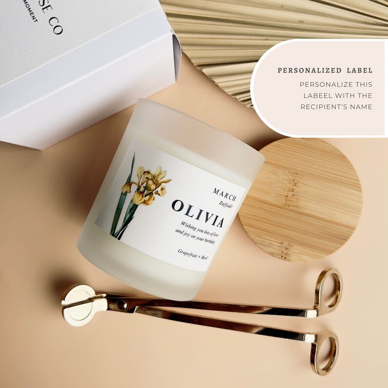 May include: A frosted glass candle jar with a white label that reads "MARCH Daffodil OLIVIA Wishing you lots of love and joy on your birthday Grapefruit + Basil". The candle is on a light brown surface with a wooden lid and gold candle wick trimmers.