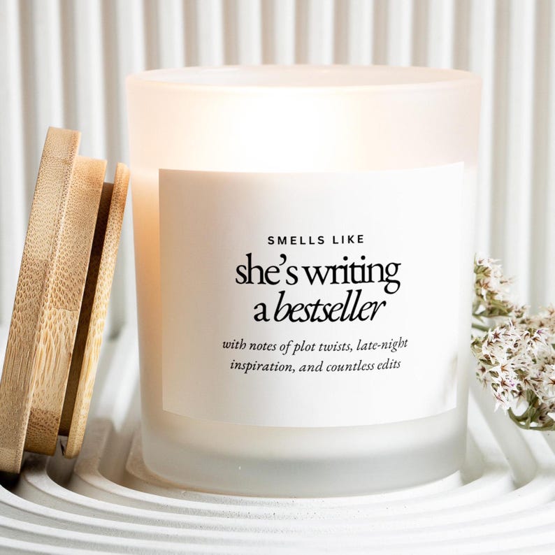 May include: A white frosted glass candle with a wooden lid, sitting on a white tray. The candle has a label that reads "Smells Like she's writing a bestseller" with additional text. A sprig of white flowers is next to the candle.