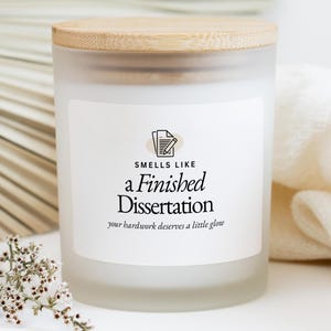 以下が含まれることがあります： 木製の蓋が付いた、すりガラスのキャンドル。ラベルには「Smells Like a Finished Dissertation」と書かれており、「your hardwork deserves a little glow」というキャッチフレーズが添えられています。キャンドルの周りには白い花と白い布があります。