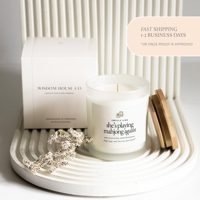 Op de afbeelding: Een witte kaars in een glazen pot met een houten deksel, naast een witte doos. De kaars heeft de tekst "Smells Like she's playing mahjong again." De doos leest "Wisdom House Co."