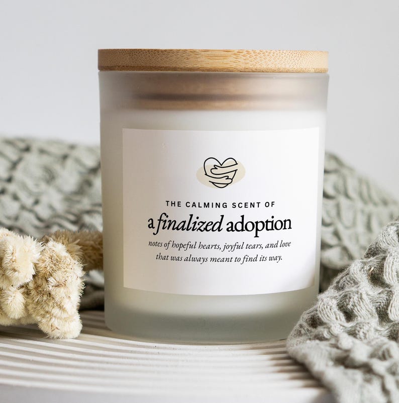 May include: A frosted glass candle jar with a wooden lid. The label reads "The Calming Scent of a finalized adoption." The text below describes the scent as notes of hopeful hearts, joyful tears, and love. The candle sits on a white surface.