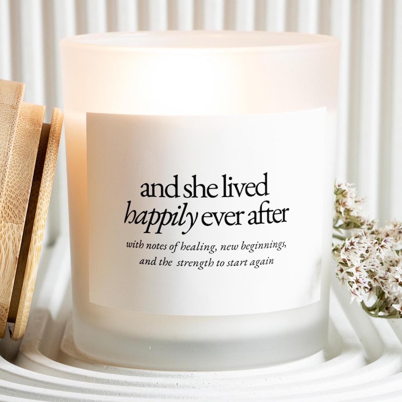 May include: A frosted glass candle with a white label featuring the text "and she lived happily ever after." The candle is in a white holder with a bamboo lid and small white flowers.