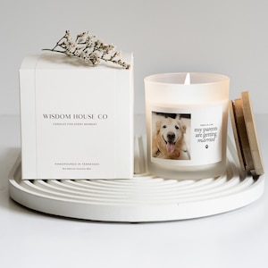 May include: A white candle in a frosted glass jar with a wooden lid, featuring a photo of a dog and the text "my parents are getting married." A white box with the text "WISDOM HOUSE CO" and "CANDLES FOR EVERY MOMENT" is next to the candle. The candle sits on a white tray.
