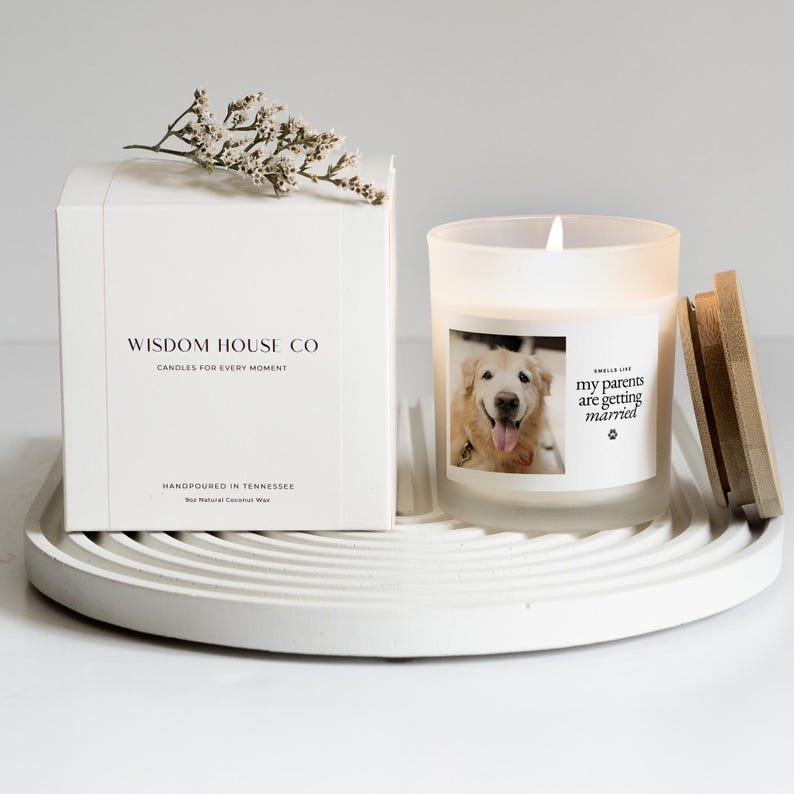 May include: A white candle in a frosted glass jar with a wooden lid, featuring a photo of a dog and the text "my parents are getting married." A white box with the text "WISDOM HOUSE CO" and "CANDLES FOR EVERY MOMENT" is next to the candle. The candle sits on a white tray.