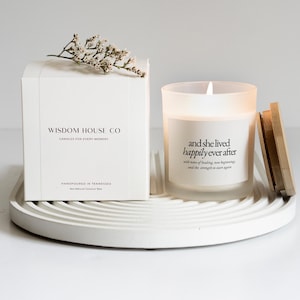 May include: A white candle in a frosted glass jar with the text "and she lived happily ever after." A wooden lid sits next to the candle. A white box with the text "Wisdom House Co" and a sprig of white flowers are also present.