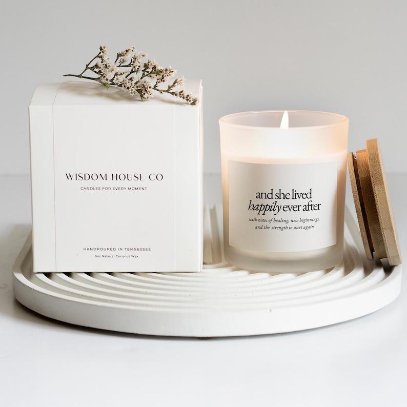 May include: A white candle in a frosted glass jar with the text "and she lived happily ever after." A wooden lid sits next to the candle. A white box with the text "Wisdom House Co" and a sprig of white flowers are also present.