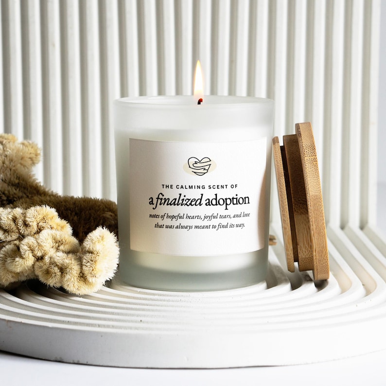 May include: A lit white candle with a wooden lid, labeled "The Calming Scent of a finalized adoption." The candle sits on a white tray with a teddy bear and a textured background. The text includes "notes of hopeful hearts, joyful tears, and love."
