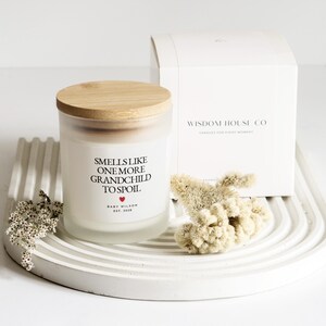 Pu&ograve; includere: Una candela bianca con coperchio in legno e la scritta "Smells Like One More Grandchild To Spoil". La candela &egrave; accanto a una scatola bianca con la scritta "Wisdom House Co." e una piccola composizione floreale su un vassoio bianco.
