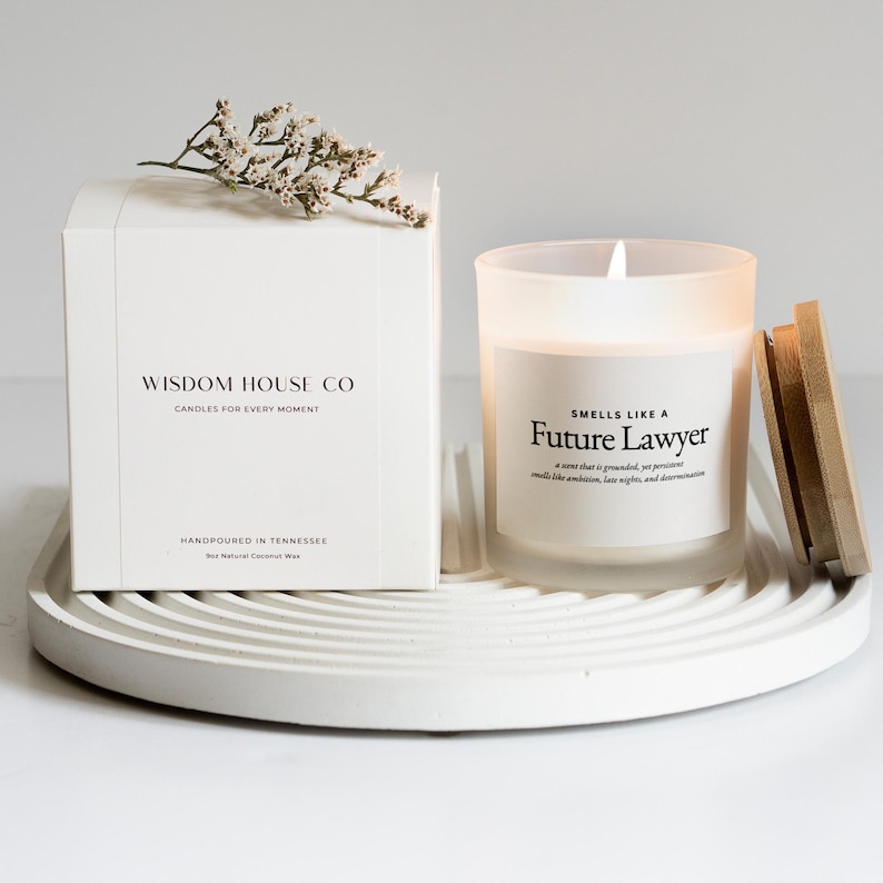 May include: A white box and a lit candle on a white tray. The box reads "WISDOM HOUSE CO CANDLES FOR EVERY MOMENT" and the candle reads "SMELLS LIKE A Future Lawyer". The candle has a wooden lid and a sprig of white flowers.
