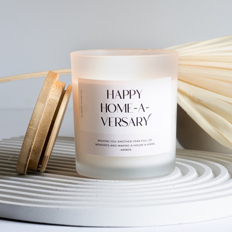 May include: A frosted glass candle with a white label that reads "HAPPY HOME-A-VERSARY" and "WISHING YOU ANOTHER YEAR FULL OF MEMORIES AND MAKING A HOUSE A HOME. -AMBER".