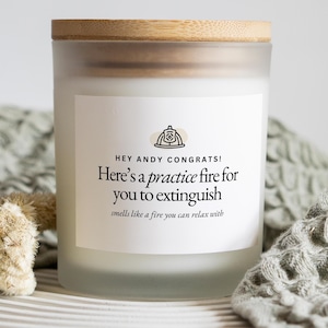 May include: A frosted glass candle with a wooden lid. The candle has a white label with the text "HEY ANDY CONGRATS! Here's a practice fire for you to extinguish smells like a fire you can relax with."