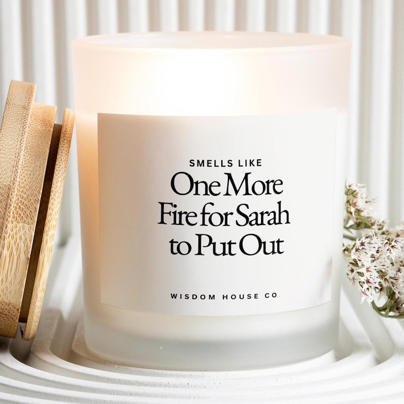 Pu&ograve; includere: Una candela in vetro smerigliato con coperchio in legno. La candela ha un'etichetta bianca con il testo "Smells Like One More Fire for Sarah to Put Out" e "Wisdom House Co." La candela &egrave; su un vassoio bianco.