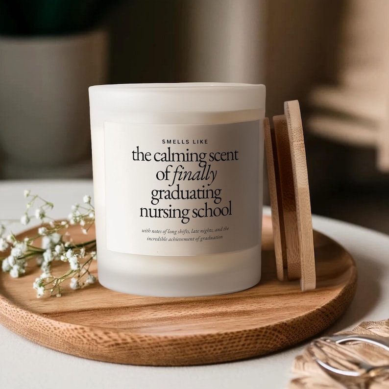 Może przedstawiać: Biała świeca z drewnianą pokrywą i drewnianą tacą. Świeca ma napis "the calming scent of finally graduating nursing school". Świeca otoczona jest białymi kwiatami.