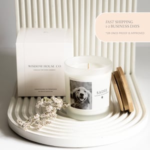 May include: A white candle in a frosted glass jar with a wooden lid. The candle has a label with a photo of a dog and the name "Sadie". A white box with the text "Wisdom House Co" is also present.