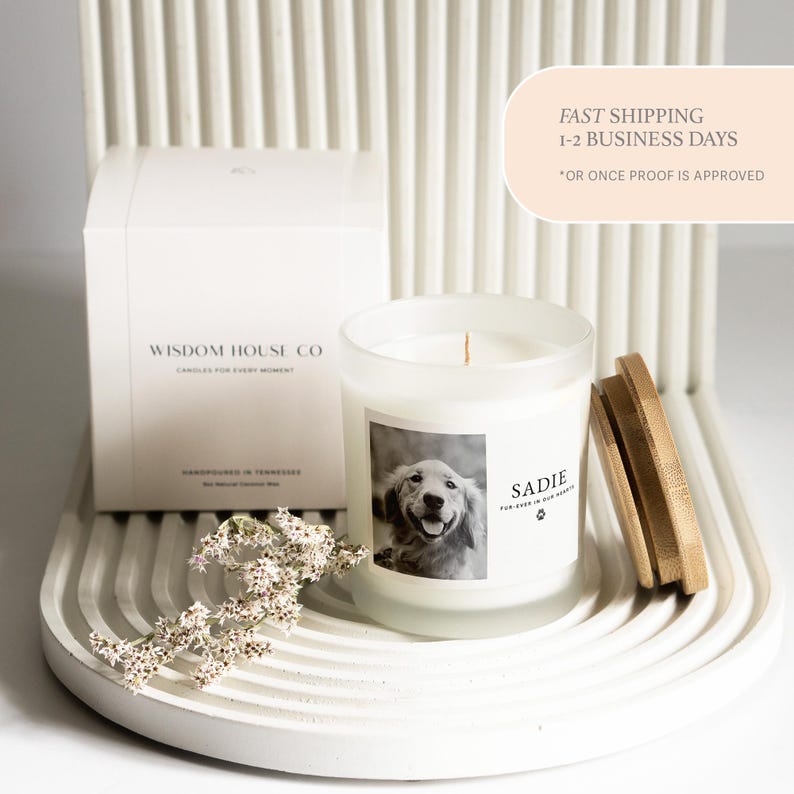 May include: A white candle in a frosted glass jar with a wooden lid. The candle has a label with a photo of a dog and the name "Sadie". A white box with the text "Wisdom House Co" is also present.