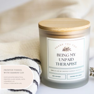May include: A frosted glass candle jar with a bamboo lid. The candle is a coconut soy candle and has a label that reads "Thank you for being my unpaid therapist. Smells like my deepest darkest secrets." The candle is 9 oz and has a burn time of 45 hours.