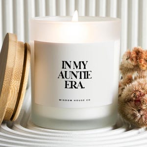 May include: A lit white candle in a frosted glass jar with a wooden lid. The label reads "IN MY AUNTIE ERA." with "WISDOM HOUSE CO" below. Dried flowers are to the right, and the candle sits on a white, ridged surface.
