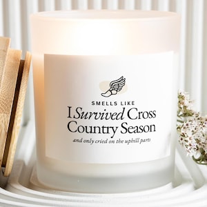 May include: Frosted glass candle with a white label. The label reads "Smells Like I Survived Cross Country Season" with a winged foot graphic. The candle is surrounded by wooden sticks and white flowers.
