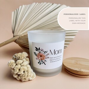 May include: A white frosted glass candle jar with a white label. The label has a floral design with a peach and pink color scheme. The text on the label reads "Mom I know you've loved me as long as I've lived but I've loved you my whole life". The candle jar has a wooden lid.