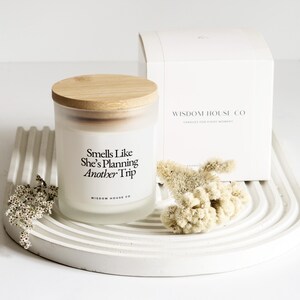 May include: A white frosted glass candle with a wooden lid, featuring the text "Smells Like She's Planning Another Trip." The candle sits on a white tray with dried flowers and a white box that reads "Wisdom House Co."