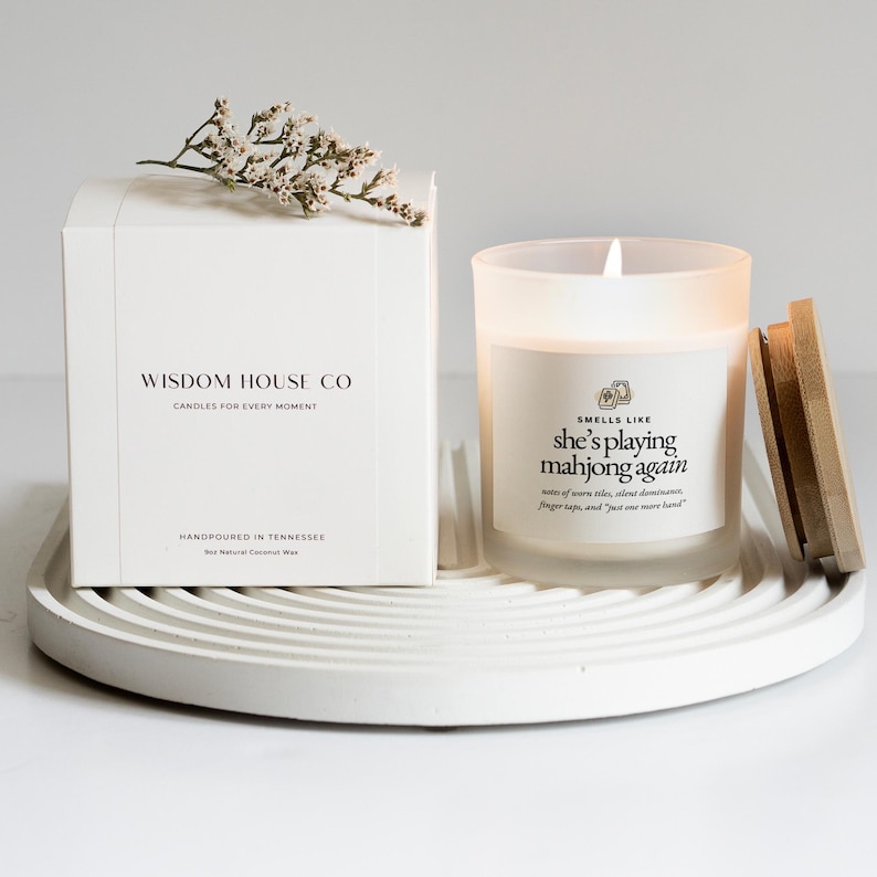 Op de afbeelding: Een witte kaars in een matglazen pot met een houten deksel, naast een witte doos. De kaars heeft de tekst "Smells like she's playing mahjong again." Op de doos staat "Wisdom House Co. Candles for every moment."
