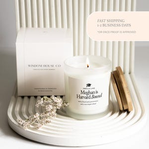 May include: A lit candle in a clear glass jar with a wooden lid. The label reads "Smells Like Meghan is Harvard Bound." A white box with the text "Wisdom House Co" is in the background. The candle sits on a white, curved tray.