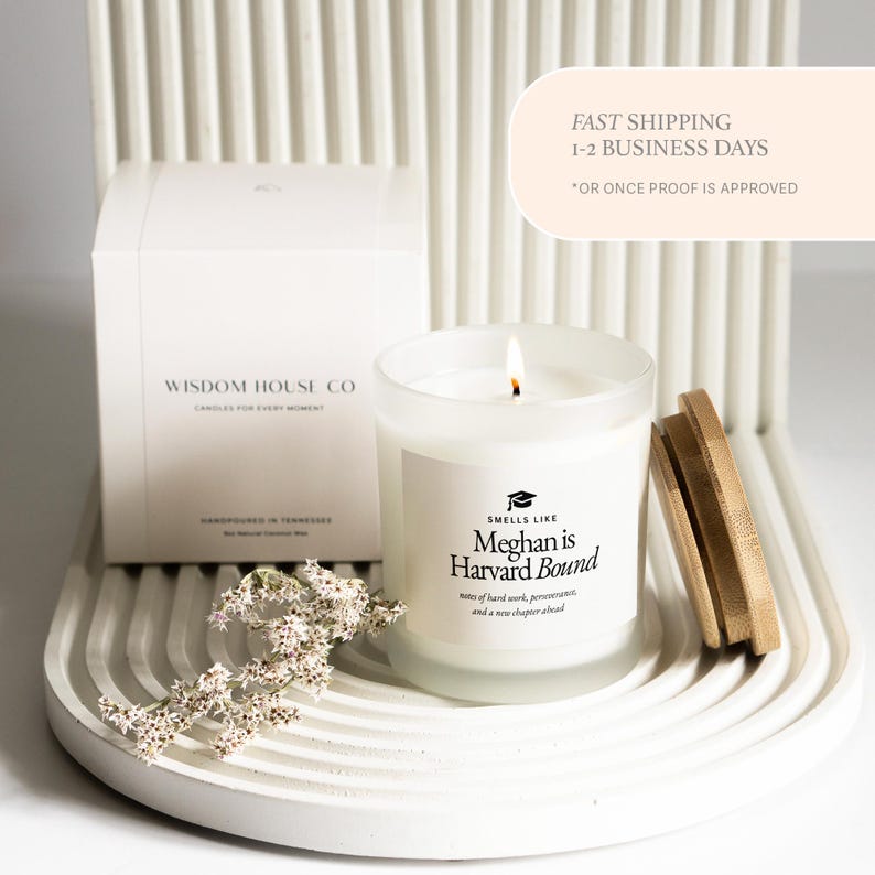 May include: A lit candle in a clear glass jar with a wooden lid. The label reads "Smells Like Meghan is Harvard Bound." A white box with the text "Wisdom House Co" is in the background. The candle sits on a white, curved tray.