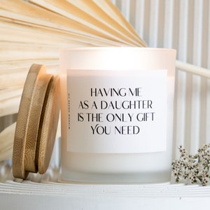 Puede incluir: Una vela de vidrio esmerilado blanco con tapa de madera, con el texto "HAVING ME AS A DAUGHTER IS THE ONLY GIFT YOU NEED". La vela está sobre una superficie blanca con un fondo decorativo.