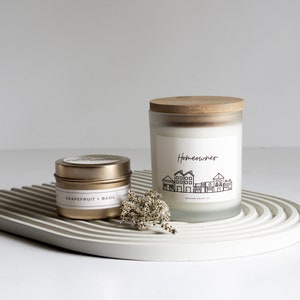Op de afbeelding: Twee kaarsen, een in een klein gouden blikje met een label met de tekst "Grapefruit + Basilicum" en een grotere kaars in een matglazen pot met een houten deksel. De grotere kaars heeft een label met de tekst "Homeowner" met een afbeelding van een rij huizen.