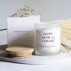 May include: A white frosted glass candle with a wooden lid. The candle label reads "Happy Home-A-Versary" and "Wishing you another year full of memories and making a house a home - Amber".