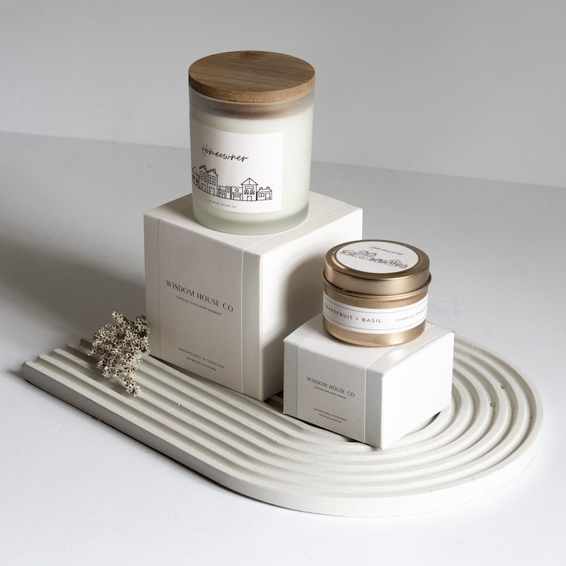 Op de afbeelding: Twee witte kaarsen met houten deksels en gouden etiketten. De grotere kaars is gelabeld "Homemaker" en de kleinere kaars is gelabeld "Grapefruit + Basilicum". Beide kaarsen staan op een wit, getextureerd oppervlak.