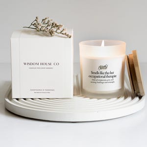 May include: A white candle and box set on a white tray. The candle has a wooden lid and text that reads "Smells like the best occupational therapist." The box reads "Wisdom House Co. Candles for every moment."