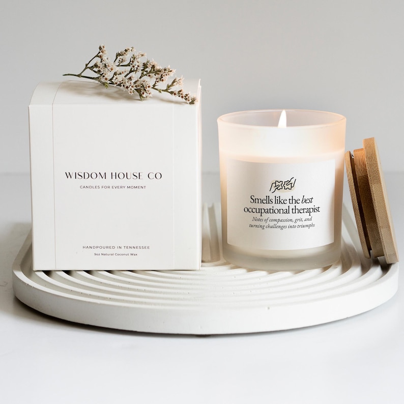 May include: A white candle and box set on a white tray. The candle has a wooden lid and text that reads "Smells like the best occupational therapist." The box reads "Wisdom House Co. Candles for every moment."
