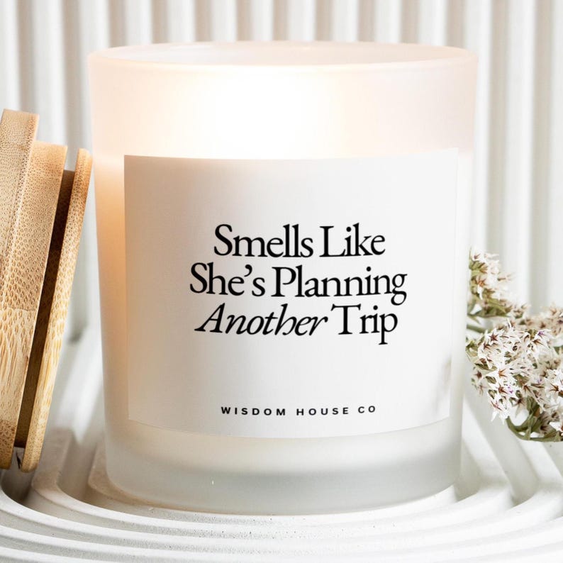 May include: Frosted glass candle with the text "Smells Like She's Planning Another Trip" and "Wisdom House Co." printed on a white label. The candle has a wooden lid and sits on a white, textured surface. Dried flowers are to the right.