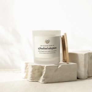 May include: A white frosted glass candle with the text "THE CALMING SCENT OF a finalized adoption" and a heart graphic. The candle sits on a white stone block with wooden sticks leaning against it. The text below reads "notes of hopeful hearts, joyful tears, and love that was always meant to find its way."
