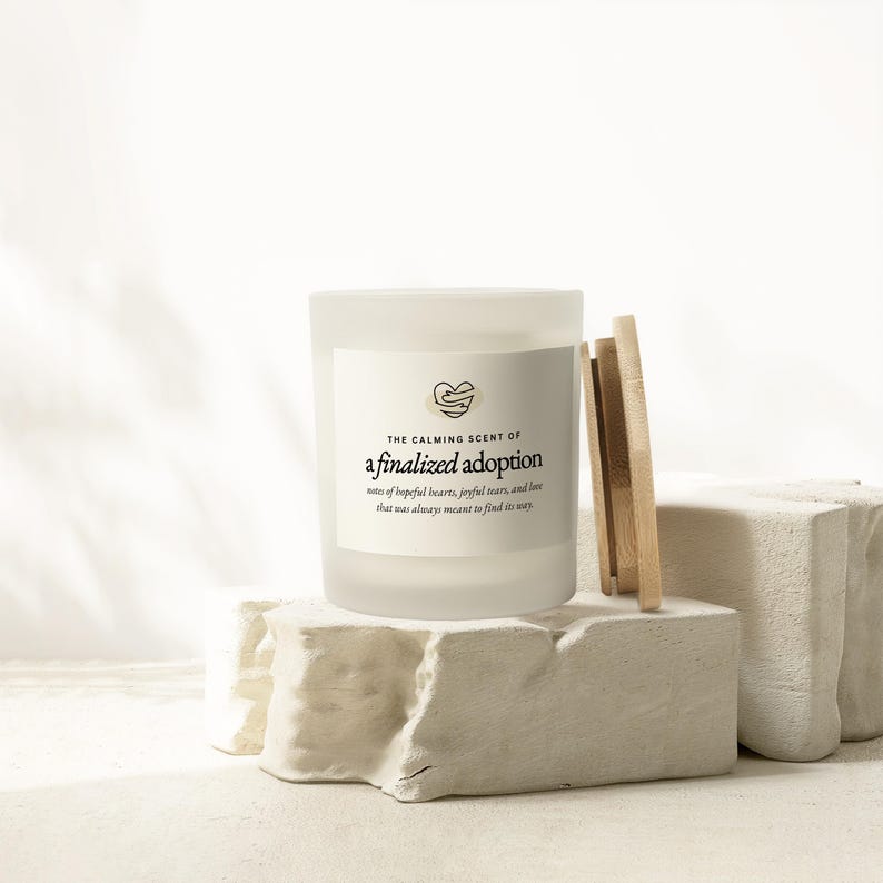 May include: A white frosted glass candle with the text "THE CALMING SCENT OF a finalized adoption" and a heart graphic. The candle sits on a white stone block with wooden sticks leaning against it. The text below reads "notes of hopeful hearts, joyful tears, and love that was always meant to find its way."