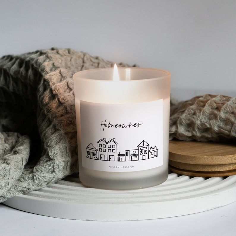 Op de afbeelding: Een witte, gematteerde glazen kaars met een zwart-wit label waarop "Homeowner" staat en een afbeelding van een rij huizen. Het label bevat ook de tekst "Wisdom House Co."