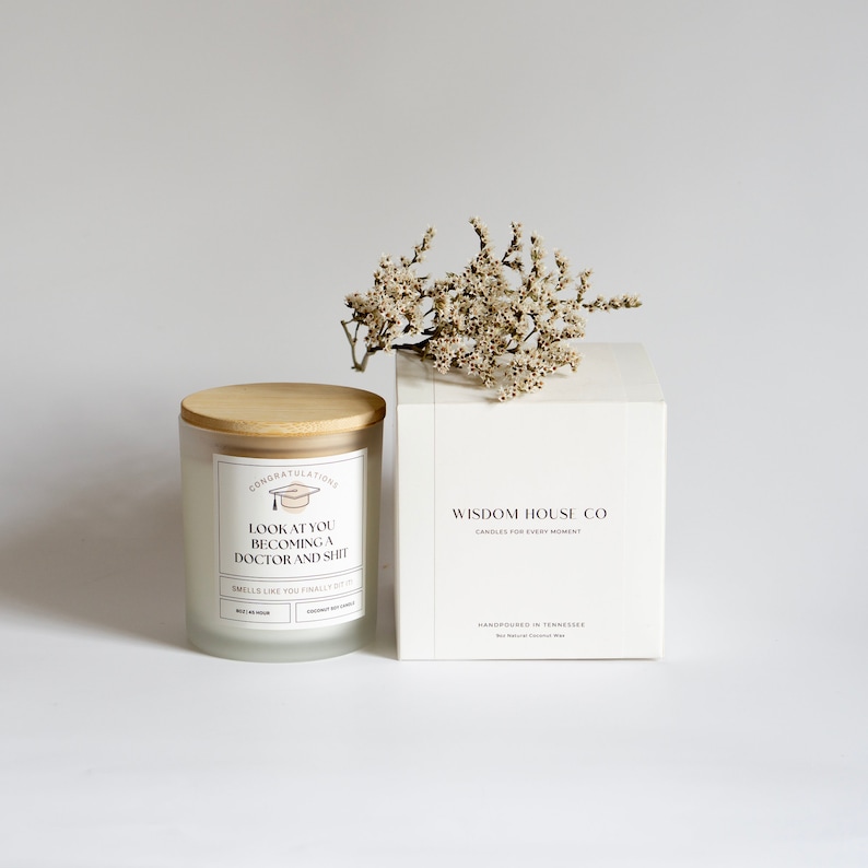Peut inclure: Une bougie en verre givr&eacute; blanc avec un couvercle en bois et un graphique de chapeau de graduation. La bougie porte le texte "Congratulations Look at you becoming a doctor and shit" et "Smells like you finally got it". La bougie est pos&eacute;e sur une surface blanche &agrave; c&ocirc;t&eacute; d'une bo&icirc;te blanche avec le texte "Wisdom House Co Candles for every moment Handpoured in Tennessee".